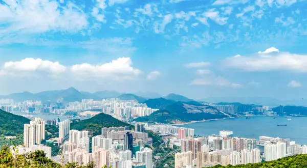 Vy över Hong Kongs tätbefolkade kustlinje med berg i bakgrunden och en blå himmel med moln.