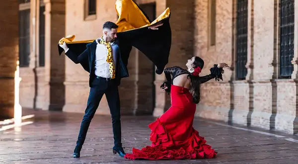 Pareja bailando flamenco con pasión. Él, con capa negra y dorada, ella, con vestido rojo voluminoso. Puro jaleo flamenco.