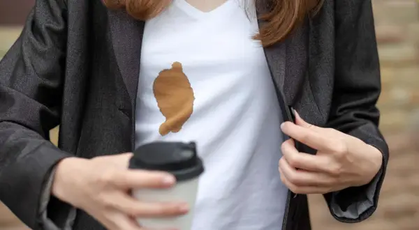 Frau mit Kaffeefleck auf weißem T-Shirt unter dunklem Blazer. Ein Becher Kaffee in der Hand. Tipps, wie man kaffeeflecken aus kleidung entfernen kann.