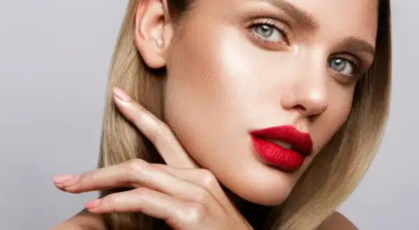 Kobieta z blond włosami i wyrazistymi, czerwonymi ustami, delikatnie dotyka policzka palcami.