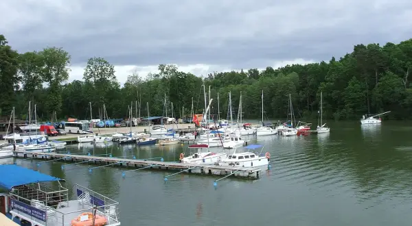 Port w Rucianem Nidzie pełen żaglówek i łodzi. W tle las i pochmurne niebo.