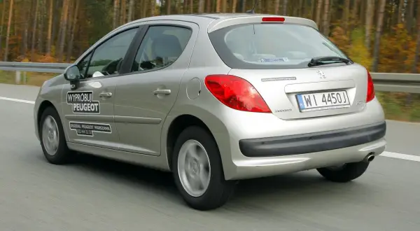 Srebrny Peugeot 207 1.4 benzyna w ruchu drogowym, z naklejkami "Wypróbuj Peugeot" i danymi oddziału.