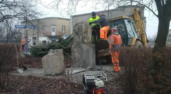 Prace przy pomniku, gdzie widać objawy zapadania się ziemi. Pracownicy w pomarańczowych strojach i koparka próbują wzmocnić podłoże.