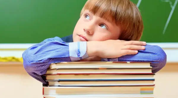 Chłopiec w niebieskiej koszuli opiera głowę na stosie książek, patrząc w górę.