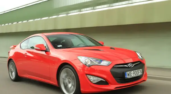 Czerwony Hyundai Genesis Coupe 3.8 V6 pędzi pod zielonym wiaduktem.