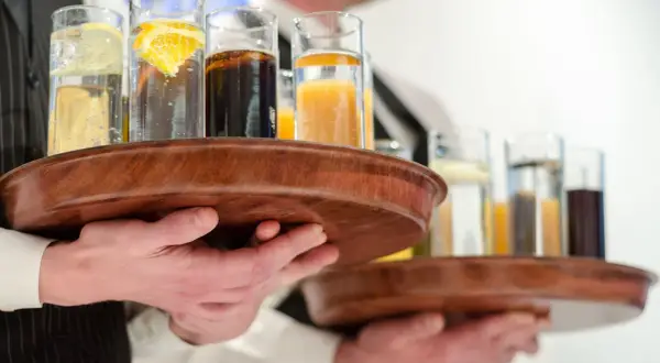 Kelnerzy niosą tace z drinkami, idealne na dzień wódki. W szklankach widać wodę z cytryną, colę i sok pomarańczowy.