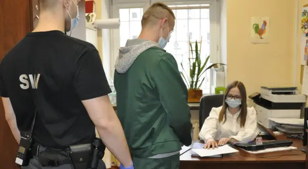 Strażnik penitencjarny w czarnej koszulce z napisem "SW" i radiotelefonem przy pasie, nadzoruje mężczyznę w zielonej bluzie, który stoi przed biurkiem urzędniczki.