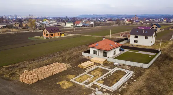 Działka inwestycyjna a budowlana: widać fundamenty domu, sterty cegieł i drewna, gotowe domy i pola uprawne w tle.