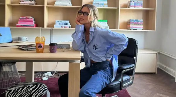 Chica con gafas sentada en una silla de oficina, luciendo un outfit oficina verano hombre: camisa azul oversize y vaqueros.