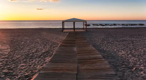 Pasarela de madera hacia una pérgola en la playa de Puerto Rey al atardecer. Sombrillas alineadas al fondo.