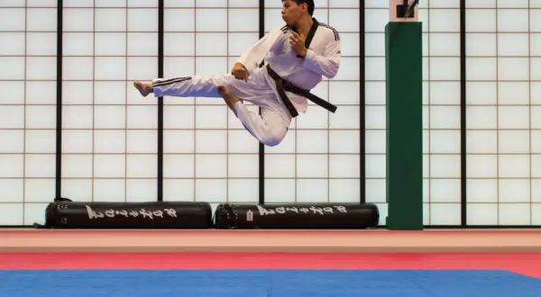 Man i vit dräkt med svart bälte utför ett högt spark i luften under en tae kwon do-träning.