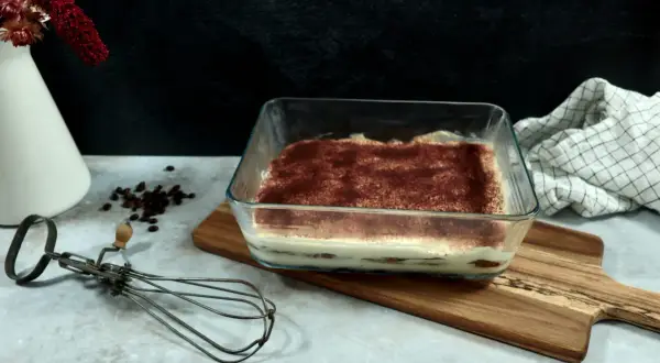 Un délicieux tiramisu 6 personnes, prêt à être dégusté, saupoudré de cacao. Un fouet ancien et des grains de café complètent la scène.