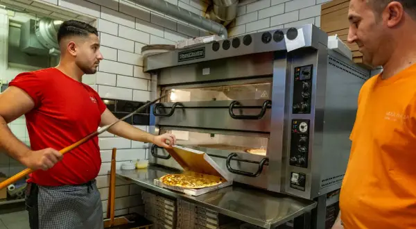 Två pizzabagare jobbar vid en stor pizzaugn. En tar ut en färdig pizza i Sverige.