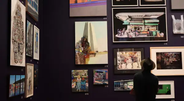 A man in a hat views diverse artworks in a gallery, showcasing vibrant cityscapes and street scenes, typical of young art galleries.