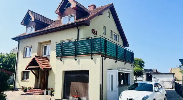 Tanie kwatery prywatne nad morzem. Dom z balkonem i białym samochodem przed posesją, otoczony zielenią.