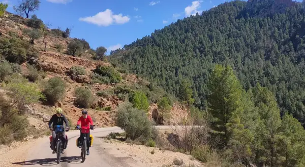 Dos ciclistas exploran rutas gravel por un camino rural rodeado de montañas boscosas y terreno rocoso bajo un cielo azul.