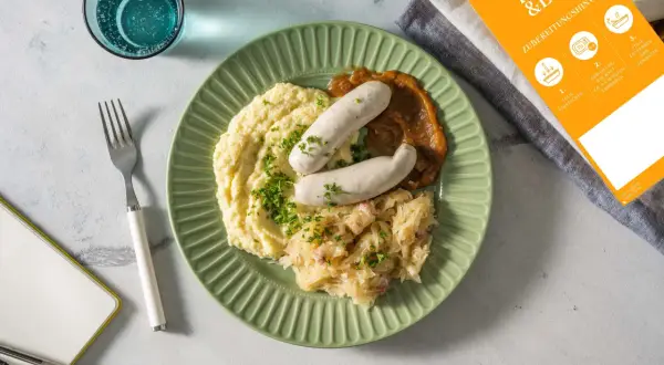 Fertig zubereitete Weißwurst mit Kartoffelpüree und Sauerkraut auf einem grünen Teller. Anleitung zum Weißwurst kochen liegt daneben.