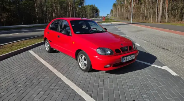 Czerwone Daewoo Lanos na parkingu z kostki brukowej, w tle las i błękitne niebo.