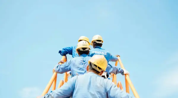 Czterech robotników w żółtych kaskach i niebieskich kombinezonach wspina się po pomarańczowych schodach. Stosują środki ochrony zbiorowej, dbając o bezpieczeństwo.