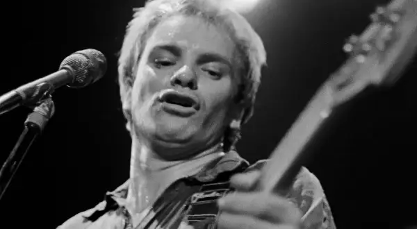 Mocno spocony muzyk, angielski singiel, śpiewa do mikrofonu, trzymając gitarę.