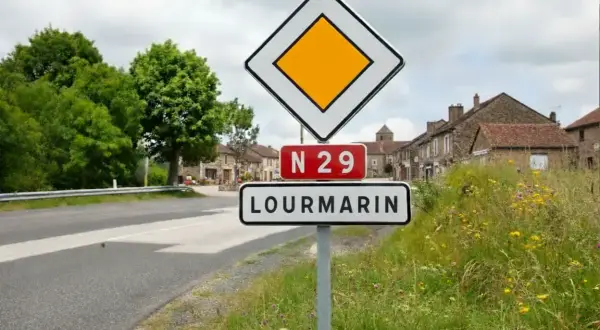 Panneau "je suis prioritaire" à l'entrée de LOURMARIN sur la N29. Le village s'étend derrière, bordé d'arbres et de maisons anciennes.