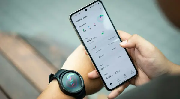 Ręka trzyma telefon Samsung z aplikacją Samsung Health, pokazującą kroki i aktywność. Zegarek na nadgarstku synchronizuje dane, pomagając jak znaleźć telefon Samsung.