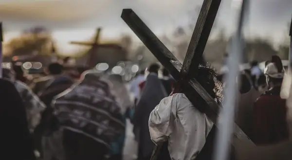 Osoba z koroną cierniową niesie krzyż, idąc w procesji. W tle widać inne krzyże i ludzi, przypominające stacje drogi krzyżowej.
