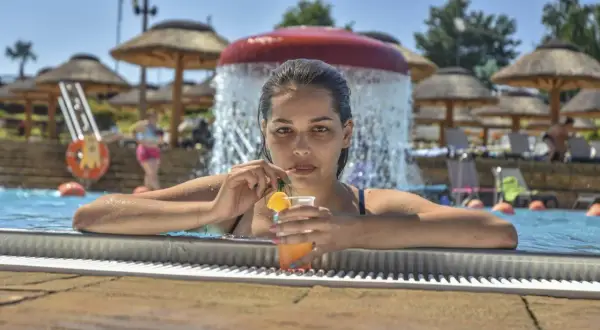 Kobieta z drinkiem w basenie, w tle wodospad i parasole. To kwintesencja wakacji all inclusive plus co to znaczy – beztroska i relaks.