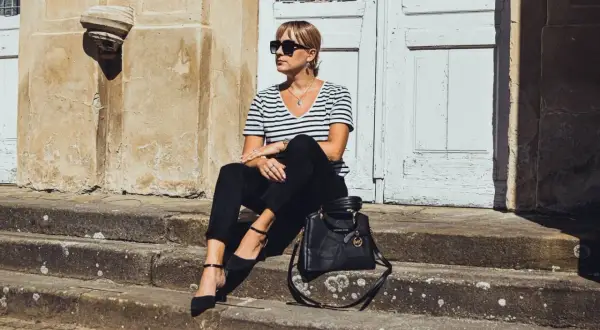 Elegancka kobieta w czarnych cygaretkach i bluzce w paski, siedząca na schodach. Stylowa torebka Michael Kors dopełnia całość.