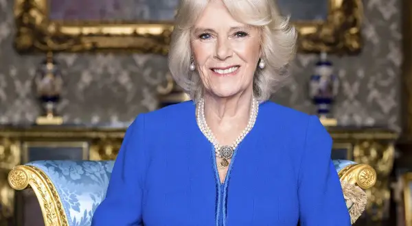 Uśmiechnięta Camilla Parker Bowles w niebieskiej sukience i perłach, siedząca na ozdobnym fotelu.