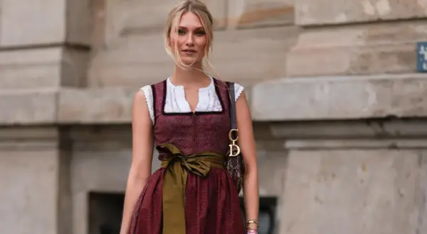 Blonde Frau in einem roten Dirndl mit weißer Bluse und einer grünen Schürze. Sie trägt eine Dior-Tasche.