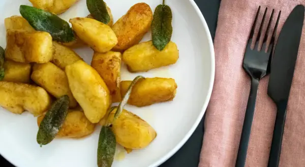 Goldbraune salbei gnocchi auf weißem Teller, garniert mit frischen Salbeiblättern. Daneben liegen schwarzes Besteck auf rosafarbenem Stoff.