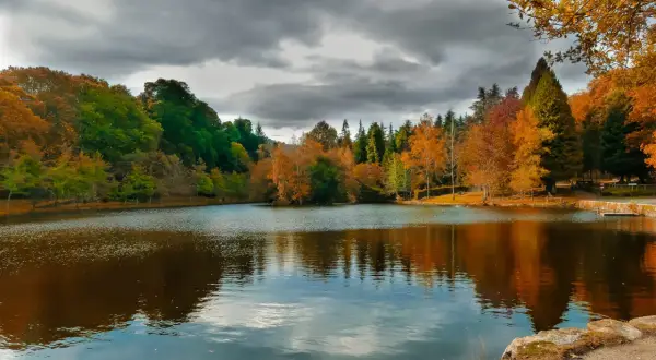 Reflejos otoñales en el lago Castiñeiras, con árboles de colores vibrantes y un cielo nublado.