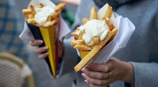 Dwie porcje frytek w papierowych rożkach, polane gęstym, białym sosem.