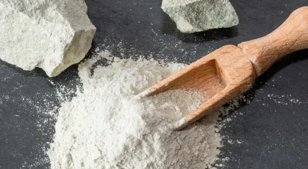 Feiner Pulver aus Magnesium-Aluminium-Silikat wird mit einer Holzschaufel auf dunklem Stein verteilt, daneben liegen zwei Brocken des Materials.