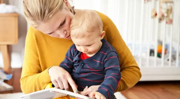 Mama uczy dziecko mówić, czytając mu książeczkę. W tle widać łóżeczko dziecięce.