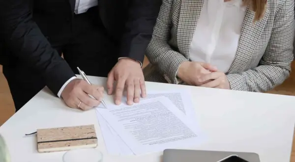 Dwie osoby przy stole podpisują dokumenty. Jedna z nich pisze piórem.