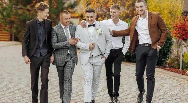 Pięciu panów w garniturach i marynarkach, świetnie ubranych na wesele, idzie razem, śmiejąc się.