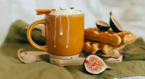 En varm kopp chai latte med vispad grädde och kanelstång, bredvid nybakade våfflor och färska fikon. Perfekt för ett mysigt chai latte recept.