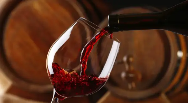 Czerwone wino leje się do kieliszka, a w tle widać drewniane beczki, które opowiadają historię wina.