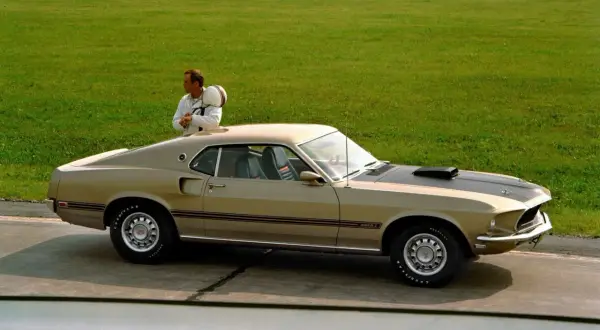 En man med hjälm lutar sig mot en guldig mustang 60 tal på en bana.