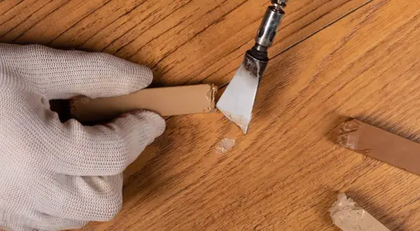 Guanto bianco rimuove residui con spatola, mostrando come togliere graffi dal parquet. Riparazione fai-da-te.