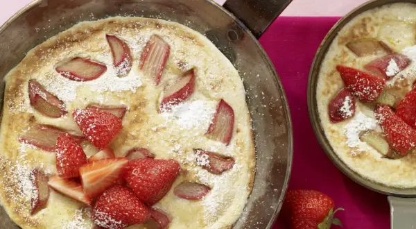 Zwei Pfannen mit frisch gebackenen Rhabarber-Pfannkuchen, garniert mit Erdbeeren und Puderzucker.