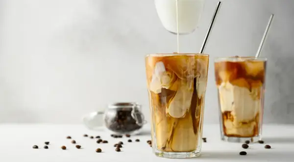 Versare latte freddo in un bicchiere di caffè ghiacciato crea un effetto marmorizzato invitante. Chicchi di caffè sparsi completano la scena.