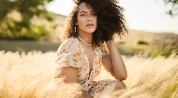 A woman with curly hair poses in a field of tall grass, her dress catching the golden hour light. This image demonstrates how to shoot with a flash for a soft, natural look.