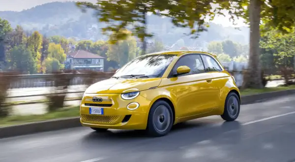 Nowy Fiat 500 w kolorze żółtym sunie po drodze, otoczony jesienną zielenią i drzewami.