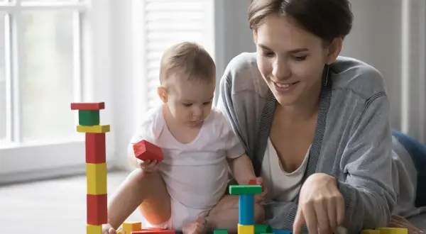 Uśmiechnięta mama bawi się klockami z dzieckiem. Zastanawiasz się, ile kosztuje opieka nad dzieckiem?