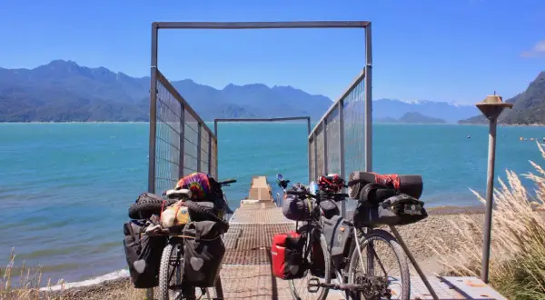 Dos bicicletas equipadas para aventura esperan junto a un muelle en la Carretera Austral. El agua turquesa y las montañas azules crean un paisaje impresionante.