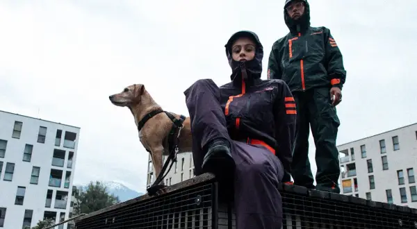 Zwei Personen in Gorpcore-Outfits und ein Hund stehen auf einem Metallgitter vor modernen Gebäuden.