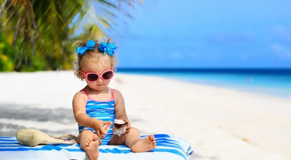 Mała dziewczynka w okularach przeciwsłonecznych i stroju kąpielowym bawi się muszlami na plaży. To idealne wakacje z niemowlakiem!
